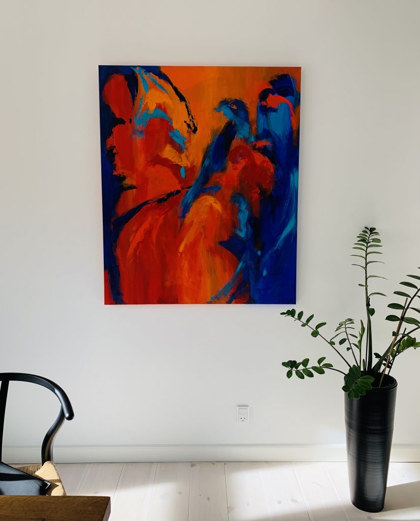 På den hvide væg i stuen hænger et stort maleri på højkant i klare farver - blå og rød - måske ser man fugle